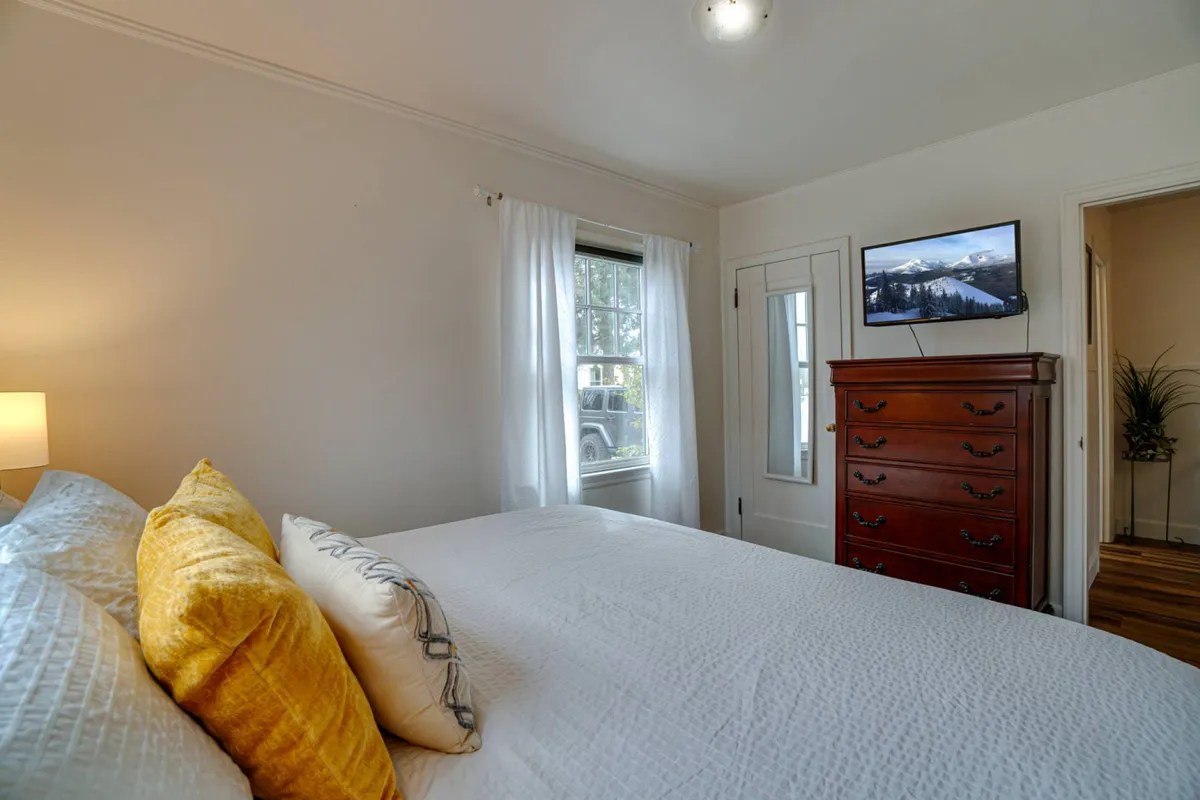 This inviting bedroom photograph showcases a bed with yellow and decorative pillows, a wooden dresser, and a TV mounted on the wall. The space feels bright and welcoming, ideal for moments of relaxation and unwinding after a busy day.