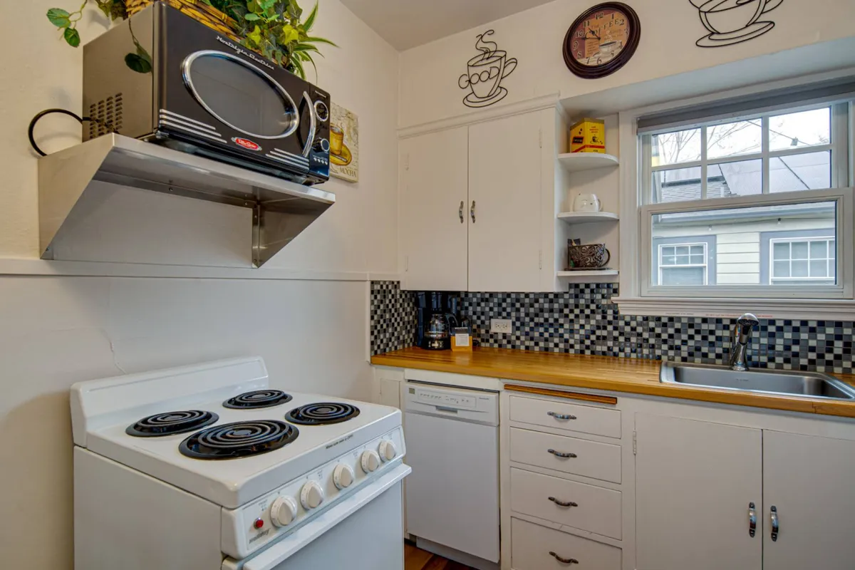 A close-up view of the kitchen area, showcasing the stove, sink, microwave, and a stylish checkerboard backsplash. Open shelves with cups and decor bring out the kitchen's practical yet homey appeal. The setup is clearly prepared for cooking and coffee making.