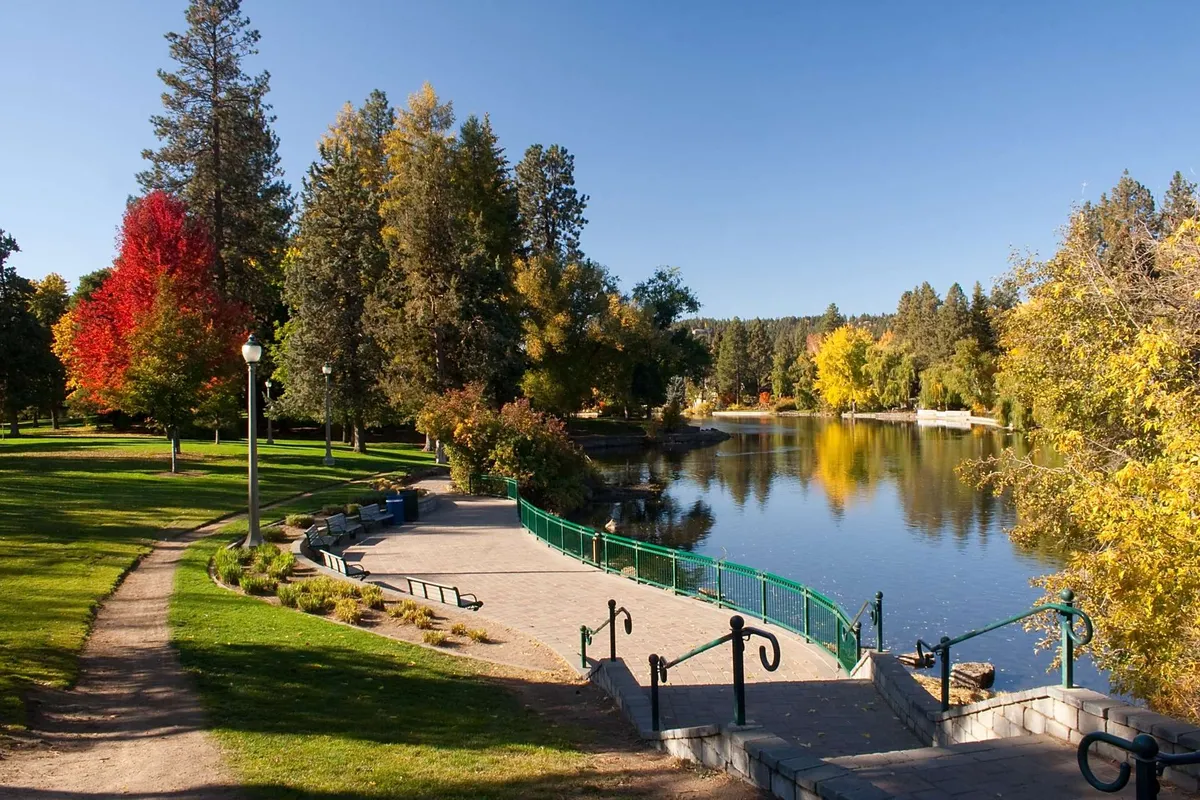 A serene view of the Deschutes River bordered by colorful trees, a winding pathway, and a charming bridge. The image captures the essence of outdoor activities like walking, biking, and nature appreciation right in the heart of town.