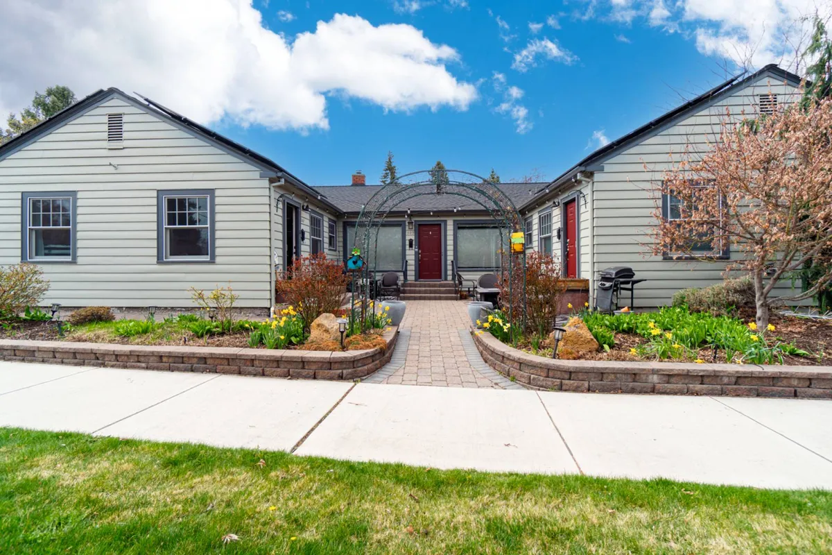 The image captures the cottage’s arched entryway surrounded by a landscaped garden and welcoming front steps. Lush greenery and spring blooms frame the entrance, hinting at the inviting environment and the walkable, charming downtown neighborhood just beyond.