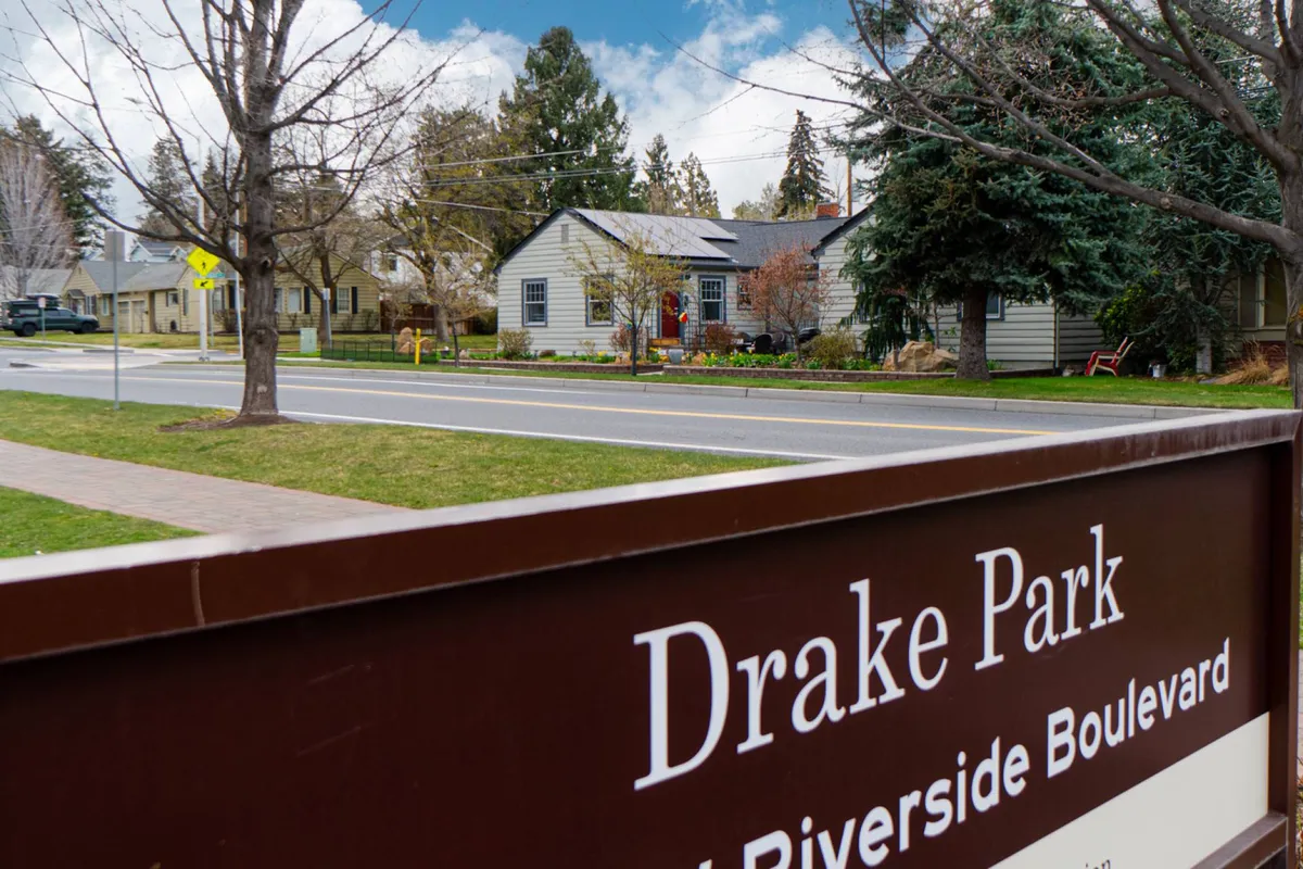 This image features the Drake Park sign surrounded by mature trees, nearby houses, and a welcoming sidewalk. The scene conveys a sense of community and accessibility, with clear visual cues of the park’s proximity to residential spaces.