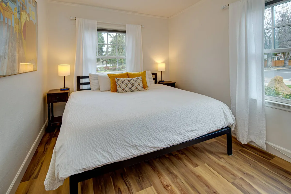 This interior image captures a plush bed with crisp linens, flanked by nightstands holding lamps, and a window with sheer curtains allowing in soft morning light. Artwork above the bed and subtle decor elements nod to a historic, tranquil ambiance.
