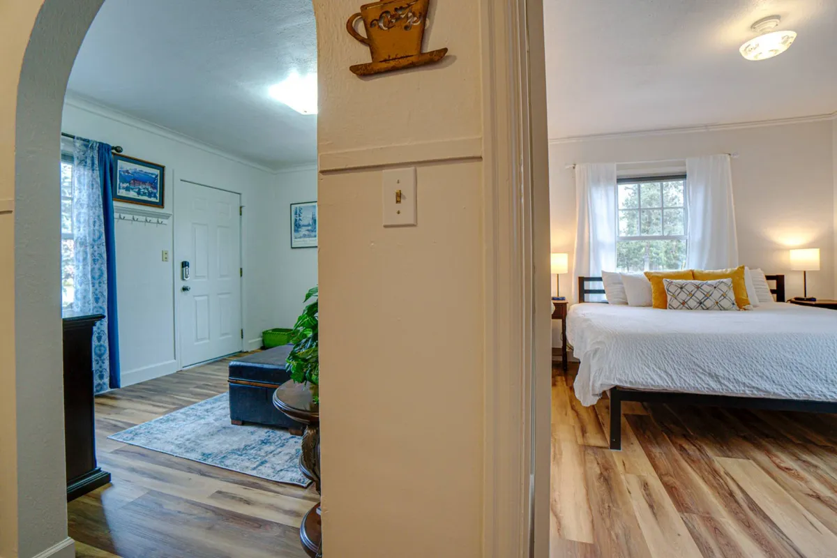 A cozy bedroom scene with a bed adorned with decorative pillows, a bright window streaming in natural light, an elegant arched doorway, and plants in the foreground. The overall mood is calm and inviting, perfect for relaxation or reading.