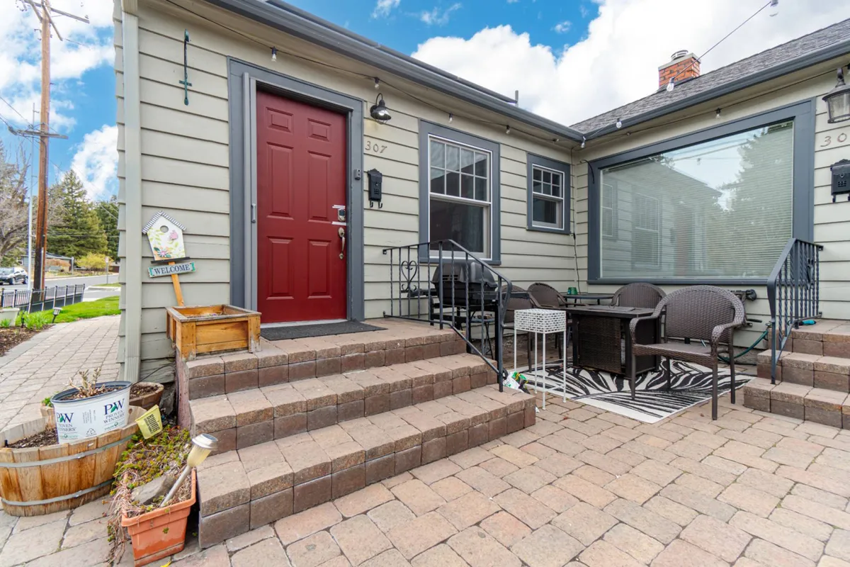 A bright, welcoming exterior patio scene with a red front door, comfortable patio seating, and a cheerful welcome sign. The setting suggests a spot ideal for relaxation, entertaining, or enjoying Bend’s urban energy just outside the doorstep.