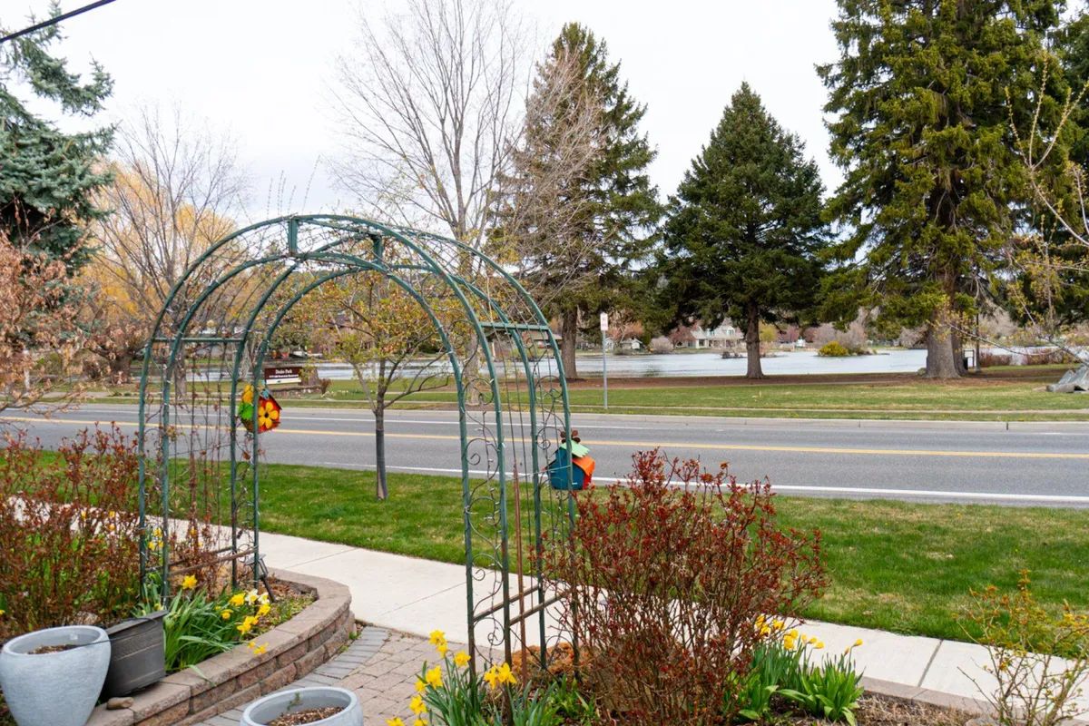 A tranquil scene with a graceful archway, lush flower beds, mature trees, a quiet road, and a peaceful lake view in the distance. The image evokes a sense of seasonal beauty and moments of relaxation amidst nature.