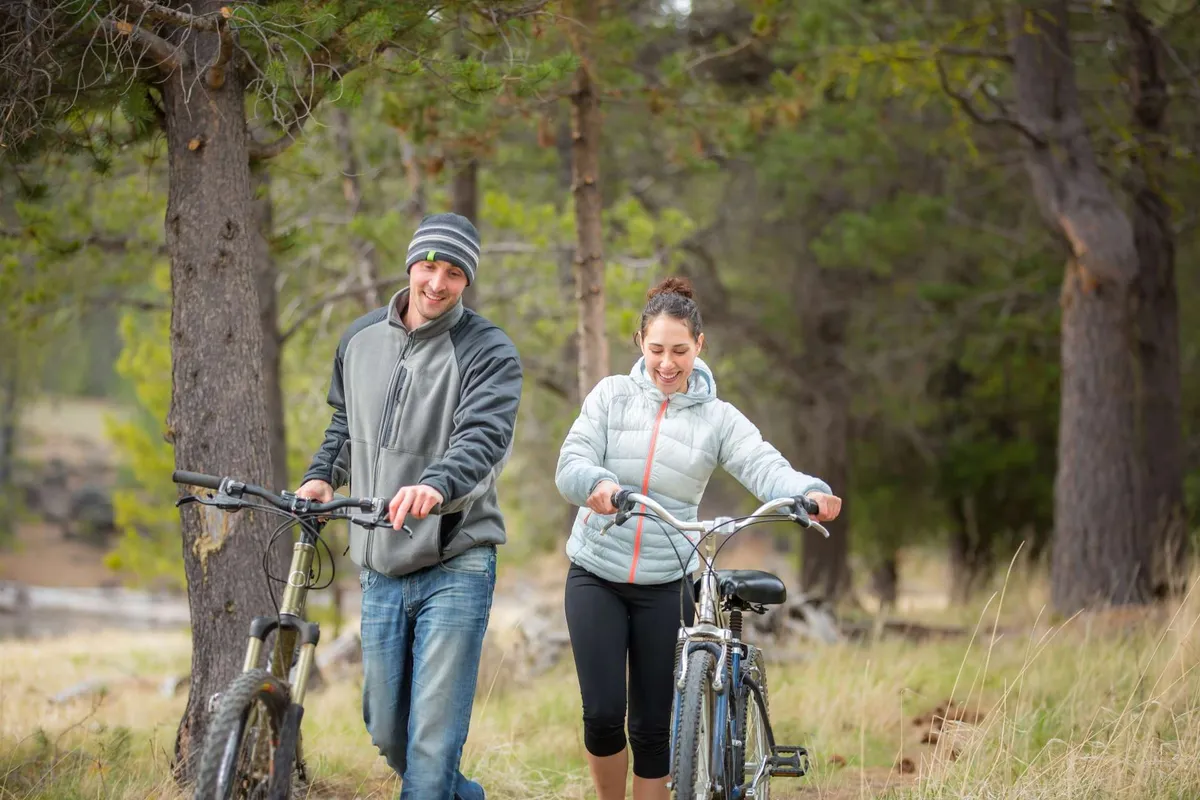 This exterior image features people walking with bicycles along a tree-lined pathway, evoking a sense of exploration and outdoor recreation. The scene is playful and relaxed, with dappled sunlight filtering through the trees and a clear, inviting path.