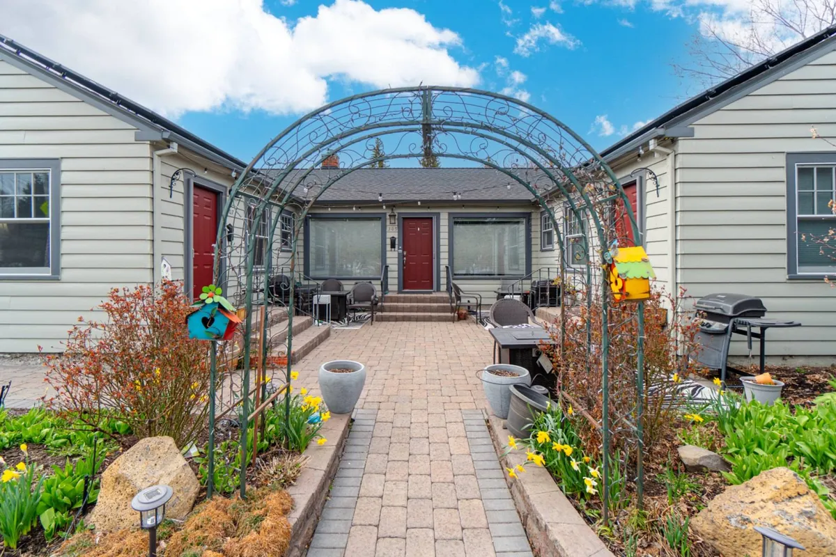 The image showcases the charming exterior of Drake Cottage 307, featuring a picturesque arched trellis draped in greenery, vibrant garden flowers, and welcoming red doors. The scene evokes a sense of warmth and hospitality, hinting at outdoor spaces perfect for relaxing or gathering.