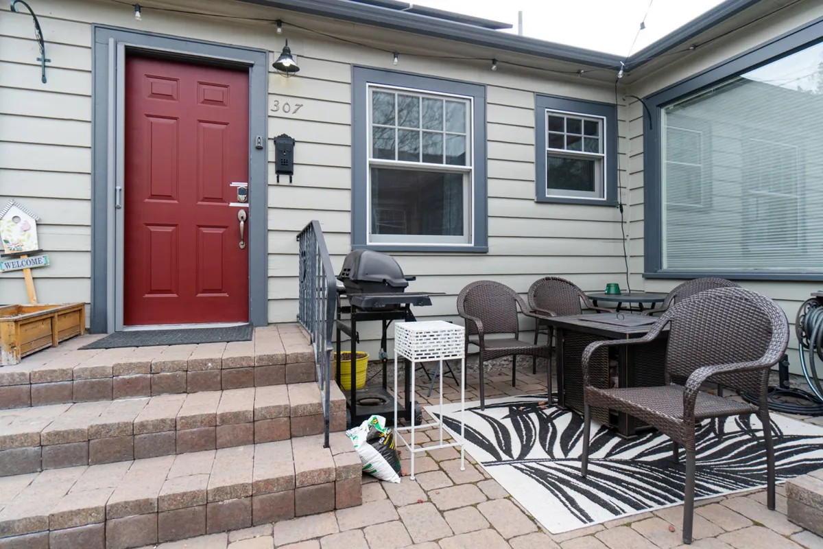 An exterior view of the home's welcoming front patio, featuring a red front door, outdoor seating area with chairs, a stylish rug, and a visible gas grill, all set for social gatherings or quiet moments. Subtle string lights overhead suggest evening ambiance.